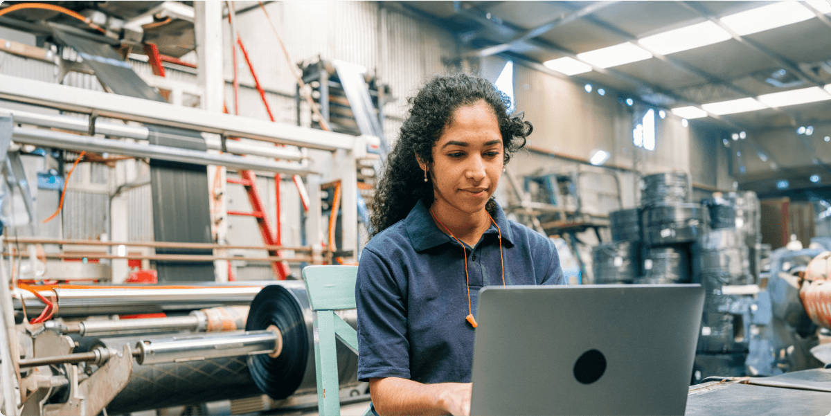 Uma mulher em uma oficina com equipamentos e ferramentas, em seu laptop.